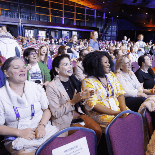A large auditorium filled with attendees seated in rows, facing a brightly lit stage.